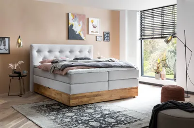 A bedroom scene with a large boxspring bed as the focal point, decorated with pillows and blankets. Artwork hangs on the wall, and a floor lamp stands near a window with blinds.
