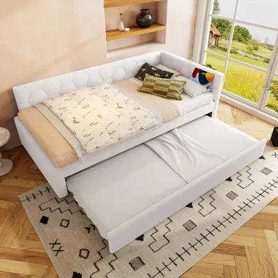 A white daybed with a pull-out trundle bed is set up in a room with a window overlooking a green landscape. Pillows and bedding are arranged on the daybed.