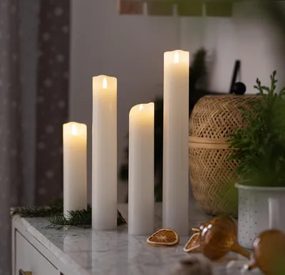 Four white LED pillar candles of varying heights are arranged on a marble surface, with a woven basket and greenery in the background.
