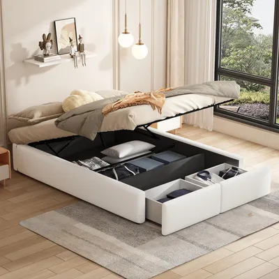 A bedroom scene with a white upholstered bed frame, its storage compartment open, revealing organized items. The bed is made with neutral-toned bedding and a throw blanket. A large window with a view of greenery is visible in the background.