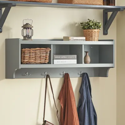 A wall-mounted gray shelf with hooks and storage cubbies, holding a basket, books, a vase, a lantern, and potted plant. Coats and a bag hang from the hooks.