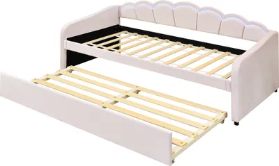 A pink upholstered daybed with a pull-out trundle bed, both showing their wooden slat bases. The daybed has a scalloped headboard with integrated LED lighting.