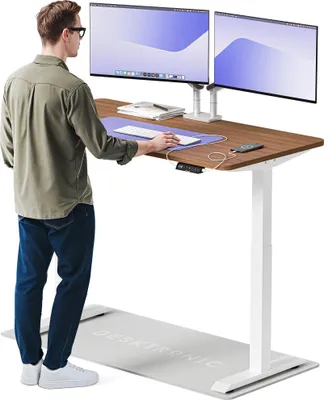 A man stands at a standing desk with two monitors, using a keyboard and mouse. A mat is on the floor in front of the desk.
