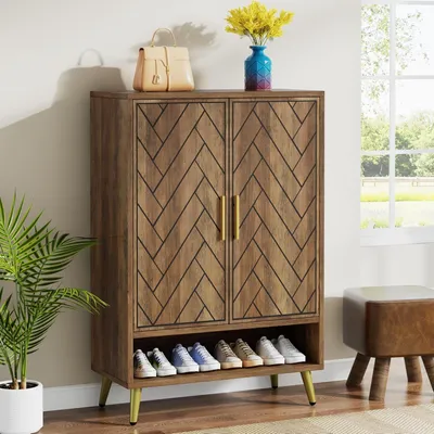 A wooden shoe cabinet with a herringbone pattern on the doors, gold handles, and gold legs. It has an open shelf at the bottom displaying several pairs of shoes. A handbag and a vase with yellow flowers are on top of the cabinet. A potted plant is to the l