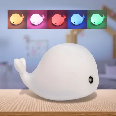 A white whale-shaped LED night light sits on a wooden surface, with a row of colored whale lights displayed above it.