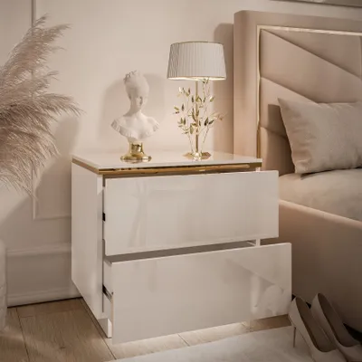 A white high-gloss nightstand with gold trim, featuring two drawers, a bust, and a decorative lamp.