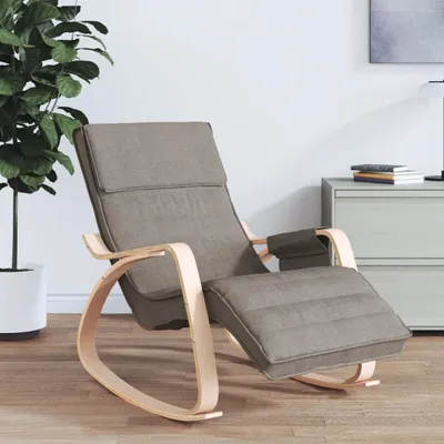 A taupe fabric rocking chair with a wooden frame is shown in a room with a plant and a dresser.