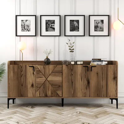 A wooden sideboard with decorative doors and black handles, placed against a white paneled wall. Above the sideboard are four framed black and white photographs. A lamp is on the left side of the sideboard, and decorative items are on top.