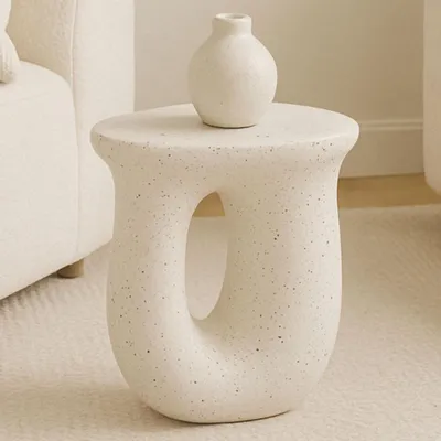 A white speckled side table with a small white vase on top, next to a textured white armchair.