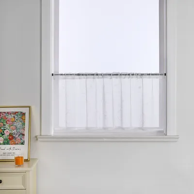 A white sheer curtain hangs on a rod in a window above a dresser with a framed picture and a candle.