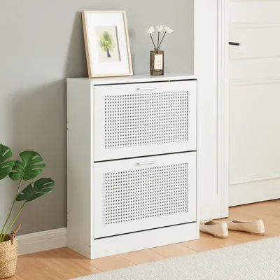 A white shoe cabinet with two flip-down doors, placed in a room next to a wall and a doorway. A framed picture and a vase with flowers are on top of the cabinet. A plant is to the left, and a pair of shoes is on the floor to the right.