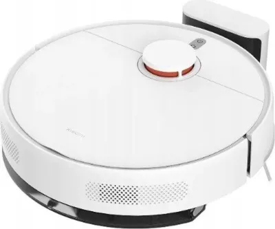 A white robot vacuum cleaner with a charging dock in the background, shown from a slightly elevated angle.