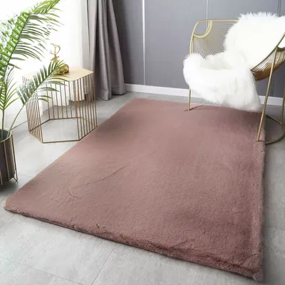 A plush rose-colored rug is laid out on a light gray floor, with a gold side table and a plant to the left, and a gold chair with a white fluffy throw to the right.