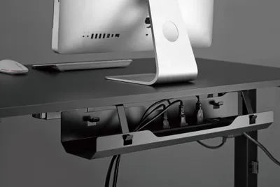 A close-up view of a cable management tray mounted under a desk, holding various cables and power strips. A computer monitor and mouse are visible on the desk surface above.