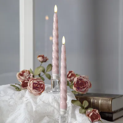 Two pink LED candles with a twisted design are displayed in clear candle holders, surrounded by dried roses and a stack of books, on a white lace tablecloth.