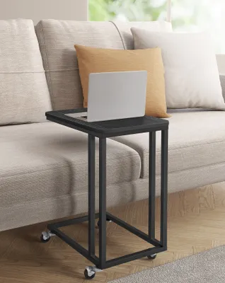 A black C-shaped side table with wheels is placed next to a beige sofa, with a laptop on top of the table. Two pillows, one orange and one white, are on the sofa.