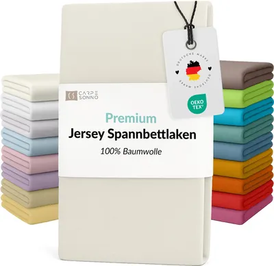 A folded cream-colored jersey fitted sheet is displayed prominently, with stacks of similarly colored sheets to the left and a vibrant stack of fitted sheets in various colors to the right. A product tag with brand and certification information is attached