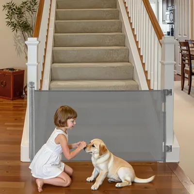 A young girl and a puppy interact at the bottom of a carpeted staircase, separated by a gray mesh baby gate.