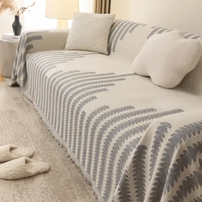 A close-up view of a sofa covered with a patterned throw blanket, with two fluffy slippers placed on the rug in front of it.