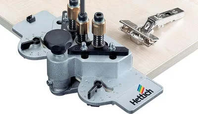 A Hettich jig for drilling holes for hinges is shown on a wooden surface, with a hinge placed next to it.