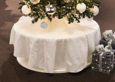 A white Christmas tree skirt is shown around the base of a decorated Christmas tree, with two gift-wrapped boxes to the right.