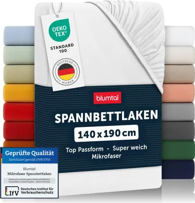 A white fitted sheet is displayed prominently, with a stack of other colored fitted sheets visible on the left and right sides. Several certification labels are attached to the sheet.