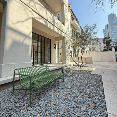 A dark green outdoor bench sits on a gravel area in front of a modern building with large glass doors and windows.
