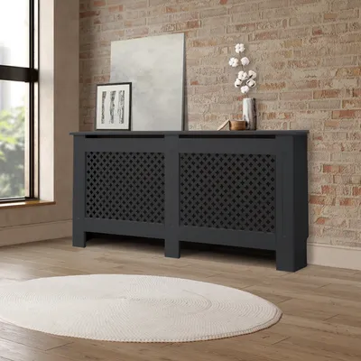 A dark gray radiator cover with a lattice pattern is placed in a room with a brick wall and a large window.