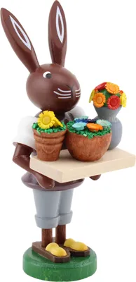 Ein fröhlicher Hase aus Holz trägt ein Tablett mit bunten Blumen in Töpfen und einer Vase und steht auf einem grünen Sockel während er ein weißes Hemd und graue Hosen trägt