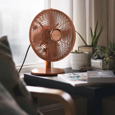 A copper-colored desk fan is placed on a windowsill next to several potted plants and books.