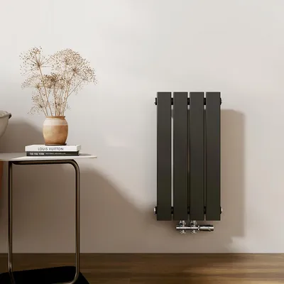 A vertical panel radiator mounted on a light gray wall, next to a small side table with books and a vase of dried flowers.