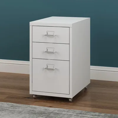 A white three-drawer filing cabinet on wheels, placed on a wooden floor in front of a teal wall.