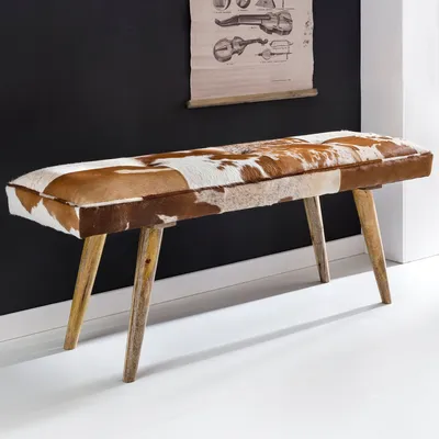 A cowhide upholstered bench with wooden legs against a black wall.