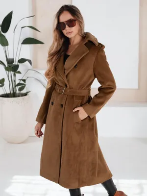 A woman wearing a brown winter coat and sunglasses stands in front of a large potted plant.