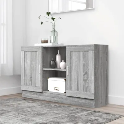 A gray wooden sideboard with two doors and shelves, decorated with vases and a box. The sideboard is placed on a gray rug in a room with a white wall and a window with white curtains.