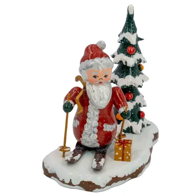 Ein fröhlicher Weihnachtsmanngehilfe in rotem Anzug steht auf Skiern im Schnee hält eine goldene Skistock in der Hand und trägt ein Geschenk während ein geschmückter Tannenbaum im Hintergrund zu sehen ist