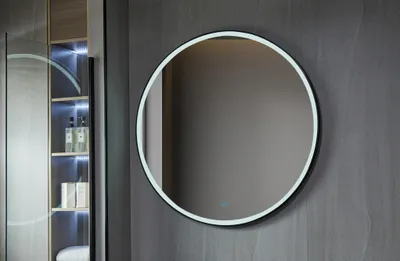 A round mirror with LED lighting is mounted on a textured gray wall, with a glimpse of a cabinet with shelves and toiletries to the left.
