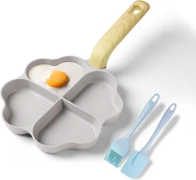 A clover-shaped frying pan with four sections, one of which contains a fried egg. Two blue silicone kitchen utensils (a brush and a spatula) are placed next to the pan.
