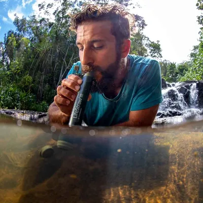 A man is drinking water from a LifeStraw filter in a natural setting with a waterfall in the background.