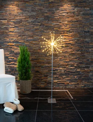 A decorative LED starburst light stands next to a potted plant and a chair, with slippers on the floor.