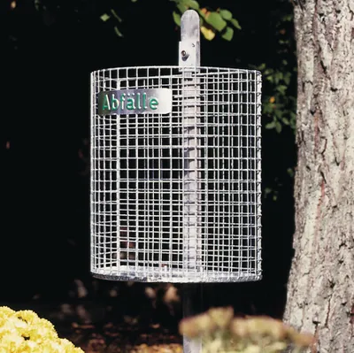 A wire mesh trash can is mounted on a pole next to a tree.