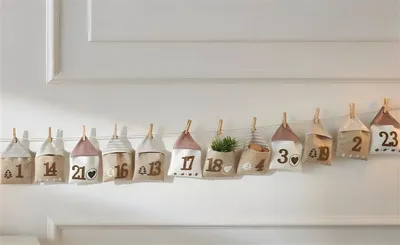 A garland of fabric advent calendar bags shaped like houses, hanging on a white wall.