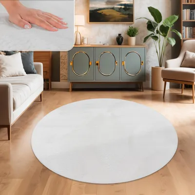 A round white shaggy rug is placed on a wooden floor in a living room setting, with a sofa, a cabinet, and a chair visible.