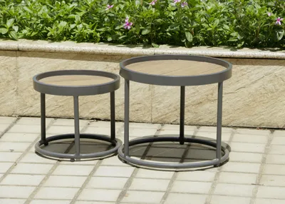 Two round side tables with wooden tops and gray metal frames are placed on a tiled patio in front of a low wall with green plants and pink flowers.
