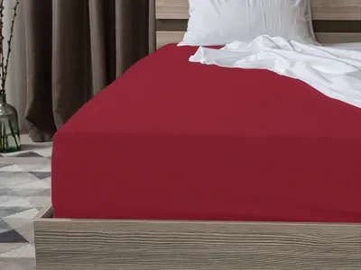 A close-up view of a red fitted sheet on a bed, with a white pillow and crumpled white sheet visible at the top.