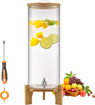 A glass beverage dispenser with a wooden stand and a metal spigot, filled with water, mango, and lime slices. A screwdriver and a ring are placed to the left, and a basket of fruit is to the right.
