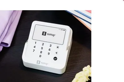 A white SumUp Solo Lite payment terminal is shown on a dark surface, with a purple cloth and a stack of books in the background.