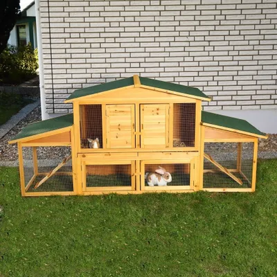 A wooden rabbit hutch with two levels and attached outdoor runs, set up on a grassy lawn in front of a brick wall.