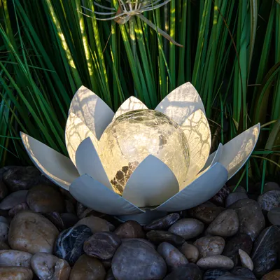A solar-powered lotus flower garden light sits on a bed of river stones, surrounded by lush green foliage.