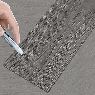 A hand is shown cutting a piece of vinyl flooring with a utility knife.